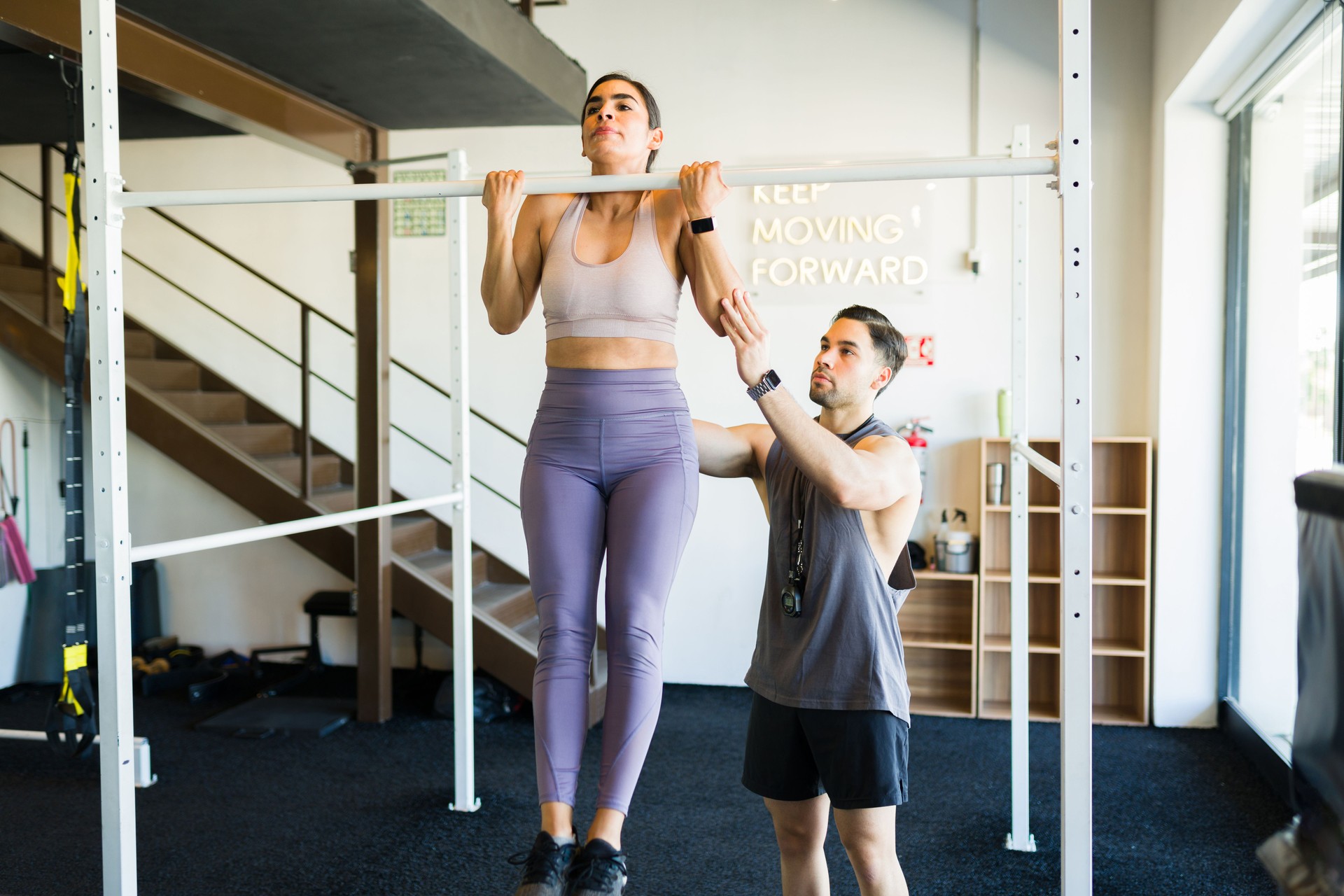 Muscular woman performing pull-ups under personal trainer supervision, demonstrating fitness commitment during intensive strength training workout